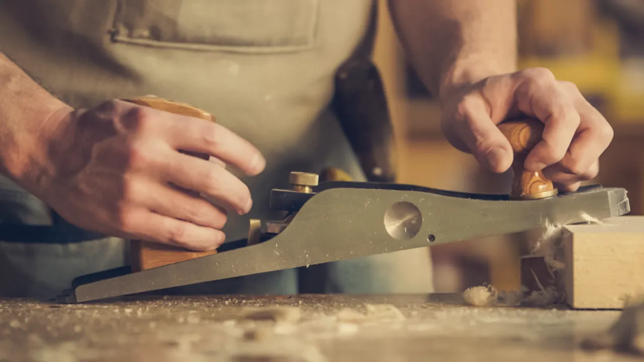A carpenter's hands guiding a steel hand plane along a board, wood shavings curling off the edge